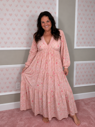 A woman wearing a long-sleeved, tiered maxi dress in a light pink floral print, standing in a room with pink heart-patterned wallpaper and a pink carpet.