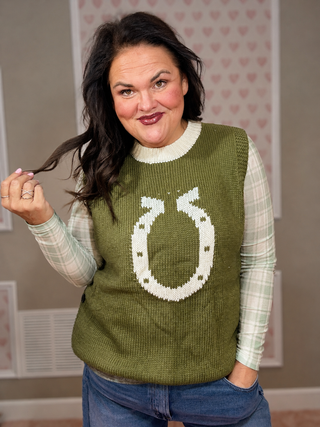 A woman wearing a green knit sweater vest with a large white horseshoe design on the front, layered over a light green and white plaid long-sleeve shirt and blue jeans.