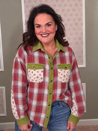 A smiling woman wearing a red, white, and blue plaid button-up shirt with olive green collar, placket, cuffs, and pocket trims, paired with blue jeans, standing in front of a wall with pink heart-patterned wallpaper.