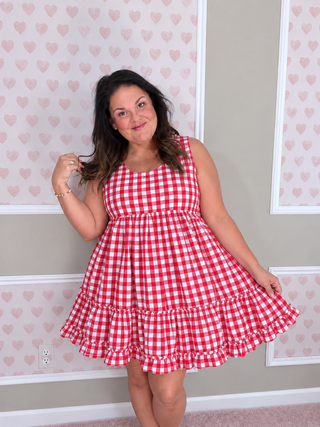 A woman modeling a sleeveless red and white gingham tiered dress in front of a wall with pink heart-patterned wallpaper.
