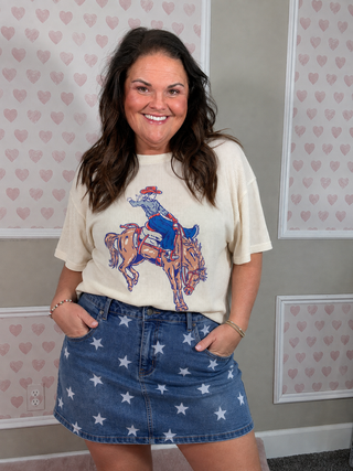 A woman wearing a cream-colored graphic t-shirt with a cowboy riding a bucking bronco, paired with a blue denim mini skirt covered in white star prints. She is standing in front of a wall with pink heart-patterned wallpaper.