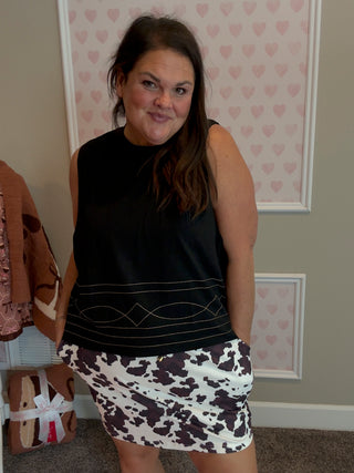 Woman wearing a black sleeveless top and patterned skirt standing in a room with heart-shaped wallpaper.