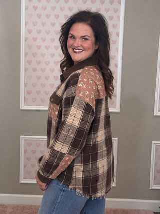 A woman wearing a brown and beige plaid patchwork jacket with floral details on the shoulders, paired with blue jeans, standing in front of a wall with pink heart-patterned panels.