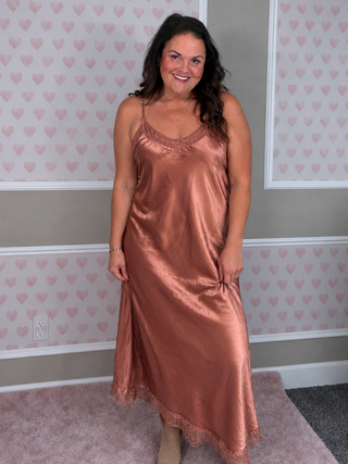 A woman wearing a satin slip dress in a dusty rose or bronze color, standing in front of a wall with pink heart-patterned wallpaper.