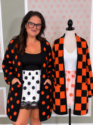 A woman wearing a black cardigan with orange polka dots stands next to a mannequin displaying a matching cardigan in a checkerboard pattern. Both cardigans are shown against a light gray wall with a pink heart patterned backdrop.