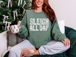 Person wearing a green sweatshirt with 'SLEIGH ALL DAY' text, holding a mug, sitting in front of a decorated Christmas tree.