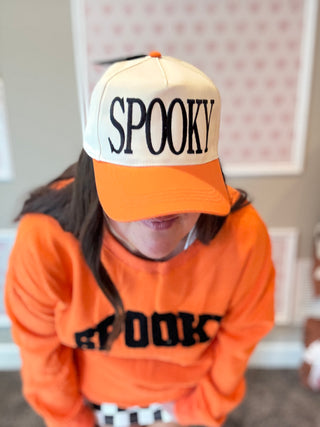 Person wearing an orange 'SPOOKY' cap and sweater in a room with a neutral background.