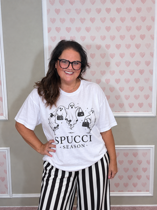 A woman wearing a white graphic t-shirt with a black ghost and handbag design and the text 'SPUCCI SEASON', paired with black and white vertically striped pants, standing in front of a pink heart-patterned wall.