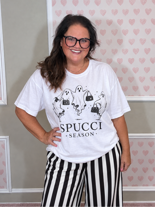 A woman wearing a white graphic t-shirt with a black ghost and handbag design and the text 'SPUCCI SEASON', paired with black and white vertically striped pants, standing in front of a pink heart-patterned wall.