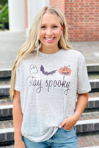 Woman wearing a 'stay spooky' t-shirt with Halloween graphics in front of a brick building.