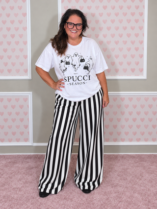 A woman modeling a white graphic t-shirt that reads 'SPUCCI SEASON' with cartoon ghosts, paired with black and white vertically striped wide-leg pants, standing against a pink heart-patterned wall.
