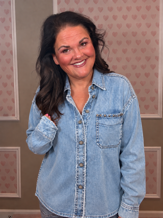 A smiling woman with long dark hair wearing a light blue denim button-up shirt, standing in front of a beige wall with a pink heart pattern.