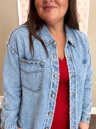 A woman wearing a light blue denim button-up shirt jacket with raw frayed edges and decorative metal studs on the chest pocket, layered over a red top.