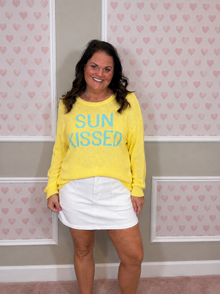 A woman wearing a bright yellow long-sleeve knit sweater with 'SUN KISSED' printed in light blue across the chest, paired with a white mini skirt, standing in front of a pink and white heart-patterned wall.