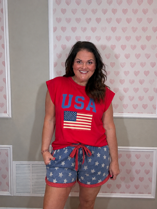 Woman wearing a red 'USA' t-shirt with an American flag and star-patterned shorts in a room with heart-shaped wallpaper.