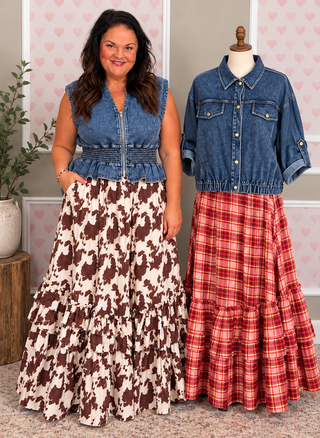 A woman stands beside a mannequin wearing two long tiered skirts with western-style denim tops. The left skirt is cream and brown cow-print, paired with a blue denim vest.
