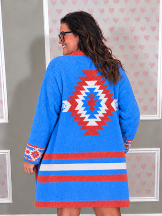 A woman shown from the back wearing a bright blue textured cardigan with a large red, white, and blue geometric diamond pattern and horizontal red and white stripes near the hem.