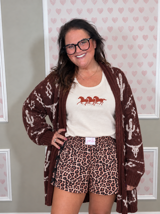 A woman modeling a brown and cream knit cardigan with cactus and western patterns, worn open over a cream graphic t-shirt and leopard-print shorts, standing against a pink heart-patterned wall.