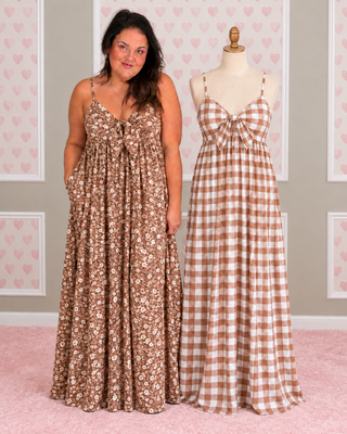 A woman stands beside a mannequin wearing two long sleeveless dresses in warm neutral tones. The dress on the mannequin is a brown and beige gingham pattern, and the dress on the woman is a matching brown gingham with a small floral print.