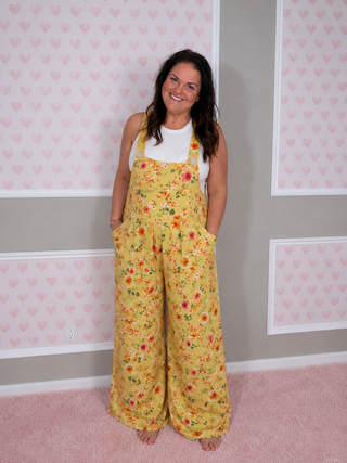 Woman wearing a yellow floral jumpsuit standing in a room with pink heart-patterned wallpaper.